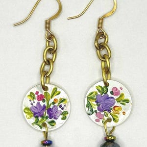 May include: Hand-painted floral earrings. Each earring features a white, circular pendant with purple, pink, and green flowers. Gold-colored chain and ear hooks connect to a faceted, dark-colored bead.