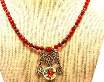 Puede incluir: Un collar con una hilera de pequeñas cuentas rojas intercaladas con espaciadores metálicos oscuros. El colgante central consta de una cuenta de cerámica pintada con flores sobre un elemento de filigrana plateada con tres dijes colgantes.