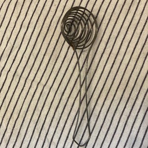 May include: A vintage wire whisk with a spiral head and a long handle. The whisk is made of metal and is resting on a striped fabric background. The whisk is designed for mixing ingredients.