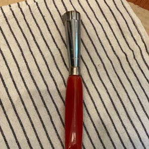 May include: A stainless steel apple corer with a red handle. The corer has a serrated cutting end and a long, slender design. The handle has a hole for hanging. The corer is resting on a striped cloth.