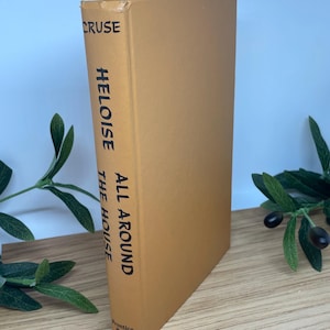 May include: A tan-covered hardcover book titled "Heloise All Around The House" by Cruse. The book's spine displays black text. The book is standing upright on a wooden surface, with green foliage in the background.