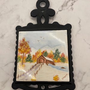 May include: Black metal trivet with a ceramic tile featuring a landscape painting of a covered bridge in autumn. The painting includes a bridge, trees, and a stream. The trivet has a decorative handle.