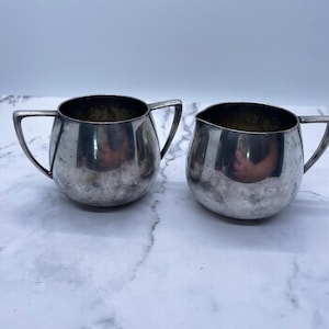 May include: Two silver-colored, small, round creamers or sugar bowls with handles. The bowls have a shiny, reflective surface and a dark interior. The handles are angular and attached to the sides of the bowls. The items are placed on a white and gray marble surface.