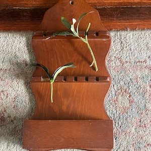 May include: A wooden wall-mounted spoon rack with a dark brown finish. The rack has two horizontal slots for spoons and a lower shelf. Two sprigs of greenery are placed in the slots. The rack is displayed against a patterned rug and a wooden wall.