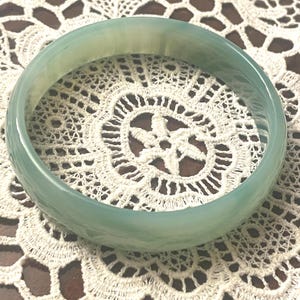 May include: A smooth, translucent pale green bangle bracelet with a polished finish. The circular jewelry piece features subtle variations in color intensity throughout its solid, continuous band structure, showcasing a soft, light-reflecting surface.