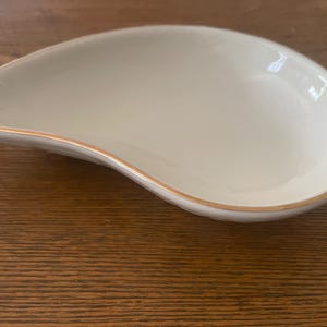 May include: A white, teardrop-shaped dish with a gold rim. The dish has a smooth, glossy surface and is likely made of ceramic or porcelain. The design is simple and elegant, suitable for serving appetizers or small portions.