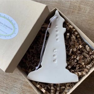 May include: A white ceramic lighthouse ornament with the word "Cape Cod" embossed on the side. The ornament is inside a cardboard box with shredded paper for cushioning. A white ribbon is attached for hanging.
