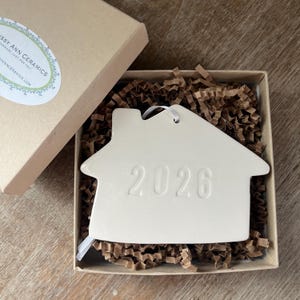 May include: A cream-colored ceramic house ornament with the year 2026 embossed on the front. The ornament is in a cardboard box with brown crinkle paper and a white ribbon. The box is partially open.