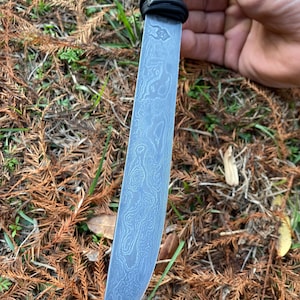 May include: A Damascus steel knife with a black wrapped handle. The blade has a unique pattern and a curved edge. The knife is held against a background of brown and green foliage.