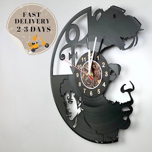 Puede incluir: Un reloj de vinilo negro con la silueta de la cara de Prince y el símbolo icónico. La esfera del reloj es un círculo más pequeño con números romanos y un diseño de engranaje.