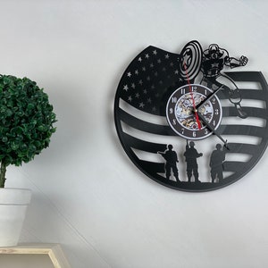 Puede incluir: Reloj de vinilo negro con una silueta de Capitán América sosteniendo su escudo, tres soldados y una bandera estadounidense. La esfera del reloj es blanca con números negros.