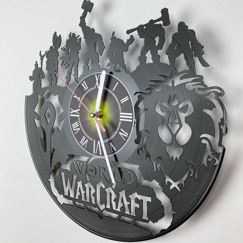 Puede incluir: Un reloj de vinilo negro con un dise&ntilde;o de World of Warcraft. La esfera del reloj es blanca con n&uacute;meros romanos negros y una aguja de reloj negra y blanca. El dise&ntilde;o incluye una cabeza de le&oacute;n y varias figuras del juego.