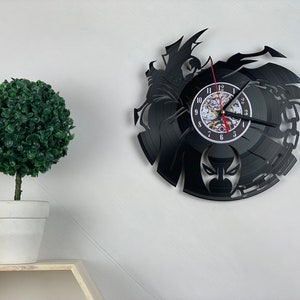 Puede incluir: Reloj de vinilo negro con una silueta de un superhéroe con máscara y cadenas. La esfera del reloj es blanca con números negros y una aguja de segundos roja.