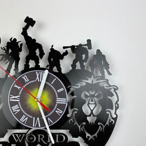 Puede incluir: Un reloj de vinilo negro con una silueta de personajes del videojuego World of Warcraft. La esfera del reloj es blanca con n&uacute;meros romanos negros y una aguja roja y blanca. El reloj est&aacute; montado sobre un fondo negro con un dise&ntilde;o estilizado de cabeza de le&oacute;n.