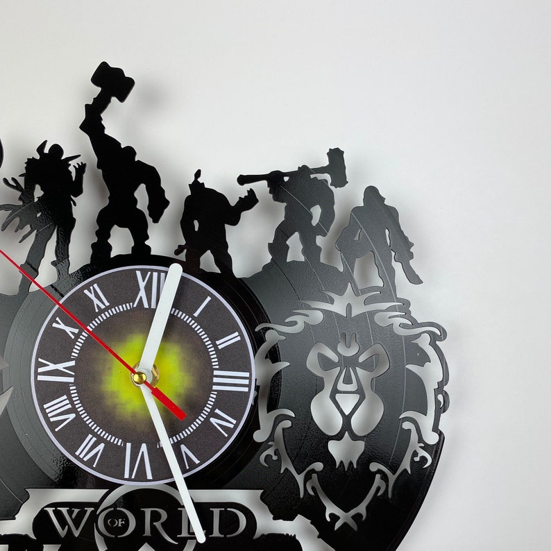 Puede incluir: Un reloj de vinilo negro con una silueta de personajes del videojuego World of Warcraft. La esfera del reloj es blanca con n&uacute;meros romanos negros y una aguja roja y blanca. El reloj est&aacute; montado sobre un fondo negro con un dise&ntilde;o estilizado de cabeza de le&oacute;n.