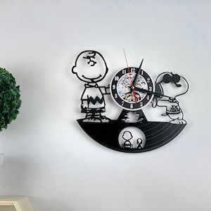 Puede incluir: Reloj de vinilo negro con los personajes de Peanuts Charlie Brown y Snoopy. La esfera del reloj es blanca con números negros y una aguja de segundos roja.