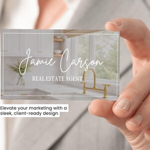 May include: A rectangular business card featuring a white border and a background image of a kitchen with white cabinets, marble countertops, and a gold faucet. The text Jamie Carson is written in a white script font, with REAL ESTATE AGENT below.