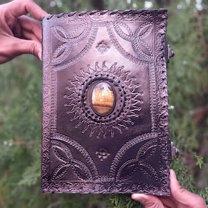 Puede incluir: Un diario de cuero marrón oscuro con intrincados diseños en relieve y una piedra preciosa de ojo de tigre engastada en el centro de la portada.
