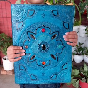 Puede incluir: Un diario de cuero turquesa con un diseño floral en relieve y detalles de piedras preciosas. La portada presenta una piedra negra central rodeada de piedras rojas, naranjas y turquesas. El diario se sostiene frente a un fondo de plantas.