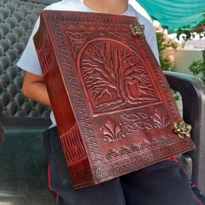 May include: A large, rectangular, leather-bound journal with a detailed tree of life design embossed on the cover. The journal is a deep reddish-brown color and features decorative metal clasps. The edges of the journal are fringed.