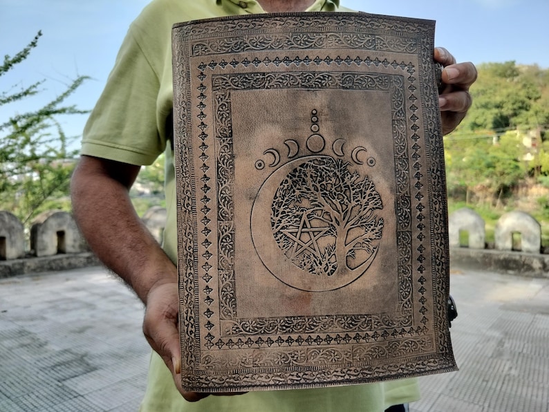 May include: A brown leather journal with an intricate design featuring a tree of life and a crescent moon. The journal has a decorative border and is perfect for writing, journaling, or sketching.