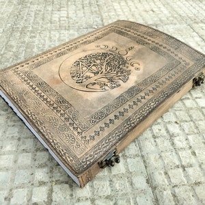 May include: A brown leather journal with an embossed tree of life design and a pentacle symbol. The journal has a decorative border and a clasp closure.