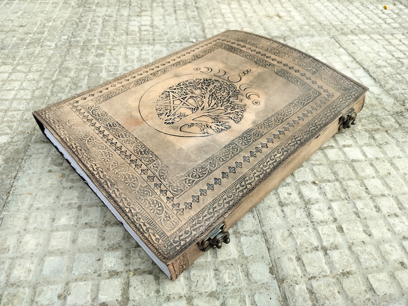 May include: A brown leather journal with an embossed tree of life design and a pentacle symbol. The journal has a decorative border and a clasp closure.
