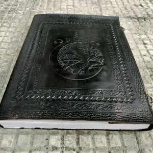 May include: A black leather-bound journal with an embossed design of a tree and a pentagram. The journal has a decorative border and a clasp.