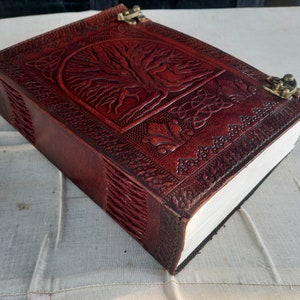 May include: A burgundy leather journal with a tree of life design embossed on the cover. The journal has a clasp closure and detailed border patterns. The pages are thick and white.