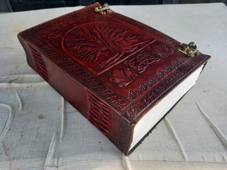 May include: A burgundy leather journal with a tree of life design embossed on the cover. The journal has a clasp closure and detailed border patterns. The pages are thick and white.