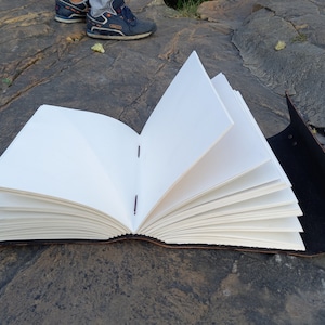 May include: A handmade journal with blank white pages, open to display the interior. The journal has a dark brown leather cover and is bound with red stitching. The pages are thick and textured. The journal is open, displaying the pages.