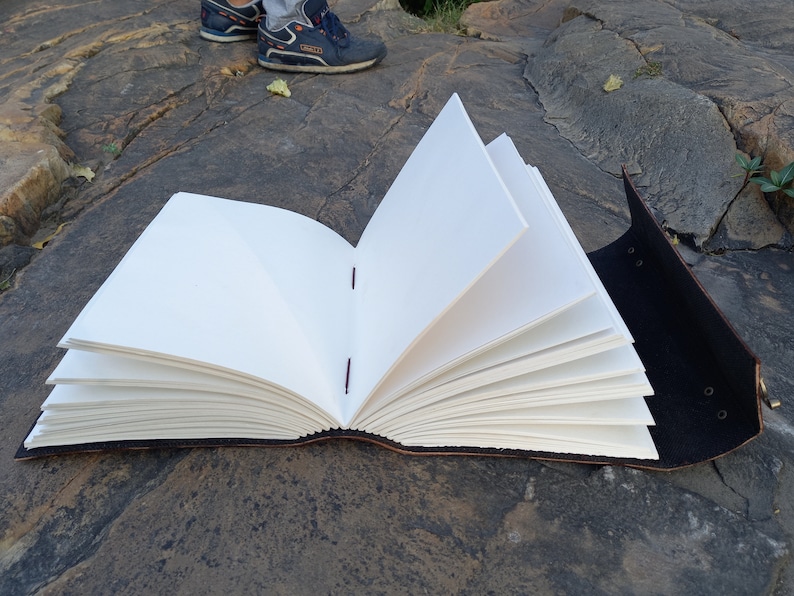May include: A handmade journal with blank white pages, open to display the interior. The journal has a dark brown leather cover and is bound with red stitching. The pages are thick and textured. The journal is open, displaying the pages.