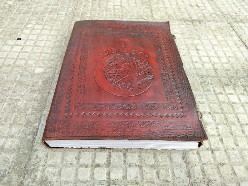 May include: A brown leather journal with a tree of life design and a pentacle symbol. The journal has a decorative border and is closed.