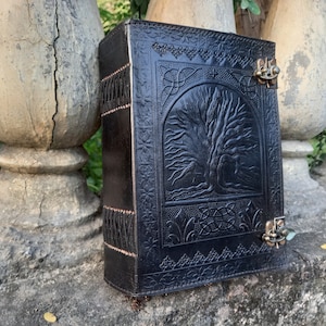 May include: Black leather journal with a tree of life design embossed on the cover. The journal has a metal clasp closure and decorative stitching along the spine. The cover features Celtic knotwork.