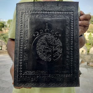 May include: Black leather journal with an embossed design of a tree of life, a pentagram, and a crescent moon. The journal has a decorative border around the edges.