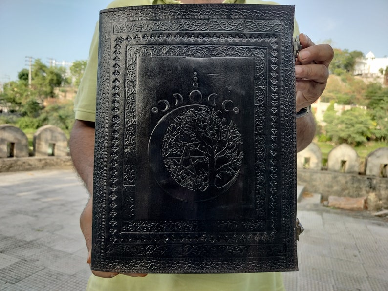 May include: Black leather journal with an embossed design of a tree of life, a pentagram, and a crescent moon. The journal has a decorative border around the edges.