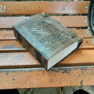 May include: A brown leather journal with a tree of life design embossed on the cover. The journal has a clasp closure and decorative floral and Celtic knot patterns. The pages are cream-coloured and the edges are dark.