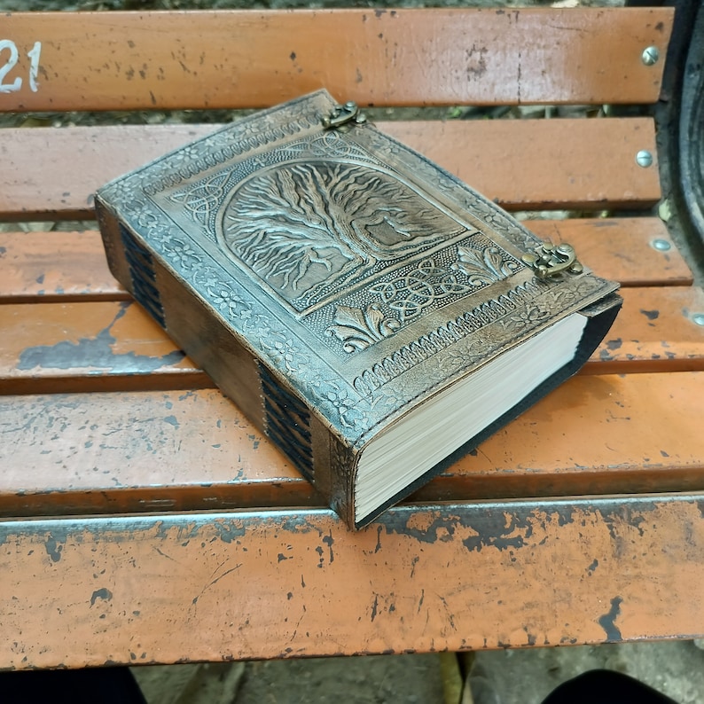 May include: A brown leather journal with a tree of life design embossed on the cover. The journal has a clasp closure and decorative floral and Celtic knot patterns. The pages are cream-coloured and the edges are dark.