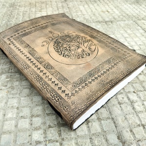 May include: A brown leather journal with an embossed tree of life design and a pentagram inside a circle. The journal has a decorative border around the edges.