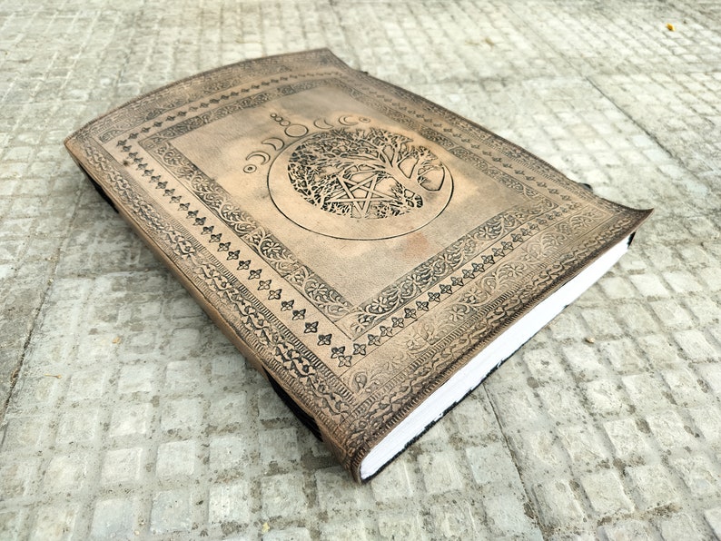 May include: A brown leather journal with an embossed tree of life design and a pentagram inside a circle. The journal has a decorative border around the edges.