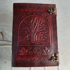 May include: A reddish-brown leather journal with an embossed tree of life design. The journal features Celtic knotwork and decorative borders. It has a metal clasp closure. Suitable for writing or sketching.