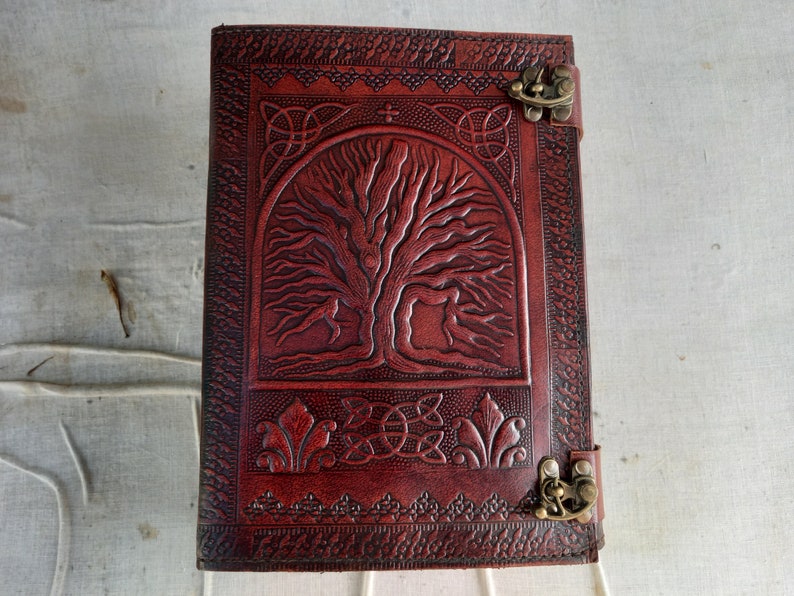 May include: A reddish-brown leather journal with an embossed tree of life design. The journal features Celtic knotwork and decorative borders. It has a metal clasp closure. Suitable for writing or sketching.