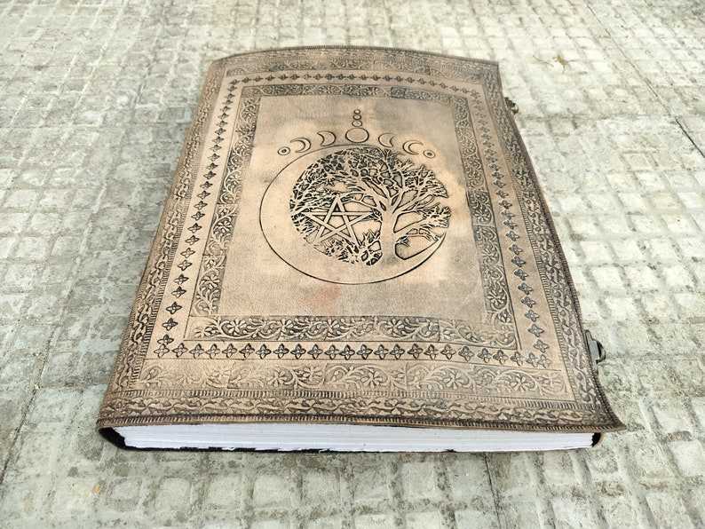 May include: A brown leather journal with a tree of life design and a pentacle inside a crescent moon. The journal has a decorative border and a clasp.