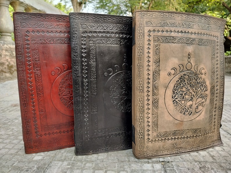 May include: Three leather-bound journals with embossed designs. The journals are red, black, and brown. Each journal features a tree of life design with a pentagram and moon phases.