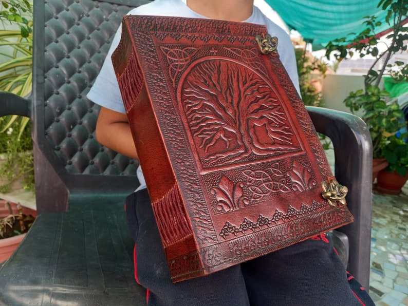 May include: A large, rectangular, leather-bound journal with a detailed tree of life design embossed on the cover. The journal is a deep reddish-brown colour and features decorative metal clasps. The edges of the journal are fringed.