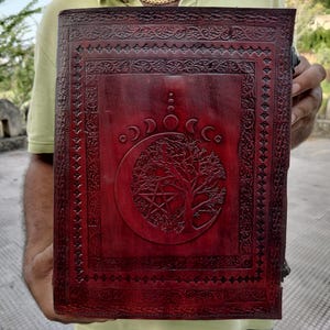 May include: A burgundy leather journal with an intricate design featuring a tree of life, a crescent moon, and a pentagram. The journal has a decorative border and is closed.