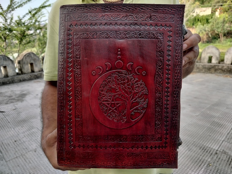 May include: A burgundy leather journal with an intricate design featuring a tree of life, a crescent moon, and a pentagram. The journal has a decorative border and is closed.