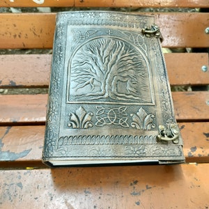 May include: A brown leather journal featuring an embossed tree of life design. The cover has decorative metal clasps and Celtic knotwork and floral patterns. The journal is closed and resting on a wooden bench.