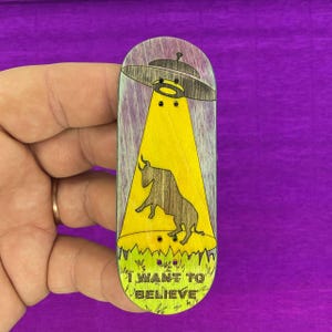 Puede incluir: Una tabla de dedo de madera con un diseño de un OVNI que proyecta un rayo de luz amarillo sobre un toro marrón. La tabla de dedo tiene el texto "I WANT TO BELIEVE" en la parte inferior, con un diseño de hierba verde. El fondo es morado.