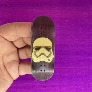 May include: A miniature wooden fingerboard with a Stormtrooper helmet design. The board is dark purple with a light tan helmet graphic. The board has three small holes for the trucks. The fingerboard is approximately 4 inches long.